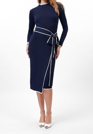 Vrouw draagt een marineblauwe geribbelde midi-jurk met lange mouwen, witte bies en een strikceintuur, gecombineerd met witte pumps met spitse neus.