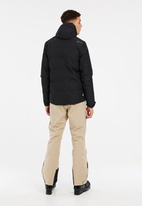 Schwarze Pufferjacke mit Kapuze, versehen mit einem gesteppten Design und elastischen Bündchen, kombiniert mit hellbeigen Hosen und schwarzen Skischuhen.