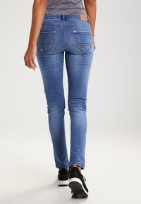 Úzké džíny v světlomodrém elastickém denimovém materiálu, s mírným vzestupem v pase, dvěma zadními kapsami a hladkým povrchem, nošené s černými teniskami.