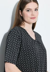 Černá blůza s geometrickým šedým potiskem, který obsahuje malé motivy jelenů. Krátké rukávy, výstřih do V a měkká látka s mírným leskem.