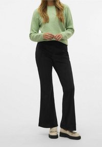 Pull vert clair à manches longues, associé à un pantalon évasé en velours côtelé noir et des chaussures beige ornées de franges décoratives.