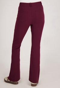 Achteraanzicht van een persoon die strakke kastanjebruine broek met achterzakken en beige schoenen draagt, staande tegen een effen achtergrond.
