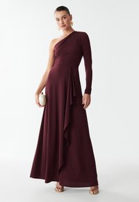 Bordeaux maxi-jurk met één schouder, een getailleerd lijfje en een gedrapeerde rok. Lange mouw aan één kant, ontspannen textuur en vloeiende silhouet.