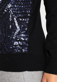 Pull en maille noire avec un motif géométrique en sequins bleu foncé et argenté sur le devant, porté avec un pantalon gris clair.