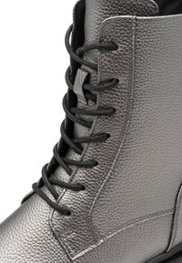 Bottine en cuir gris métallique avec une finition texturée, lacets noirs et semelle lisse. Présente une couture le long du côté.