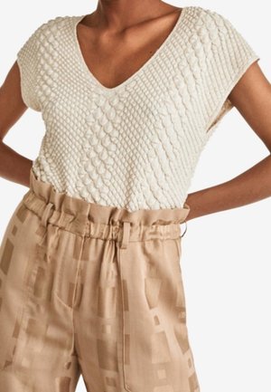Vrouw die een gestructureerde witte korte mouwen V-hals top draagt, ingestopt in beige hoge taillebroeken met een geplooide paperbag taille en geometrisch patroon.