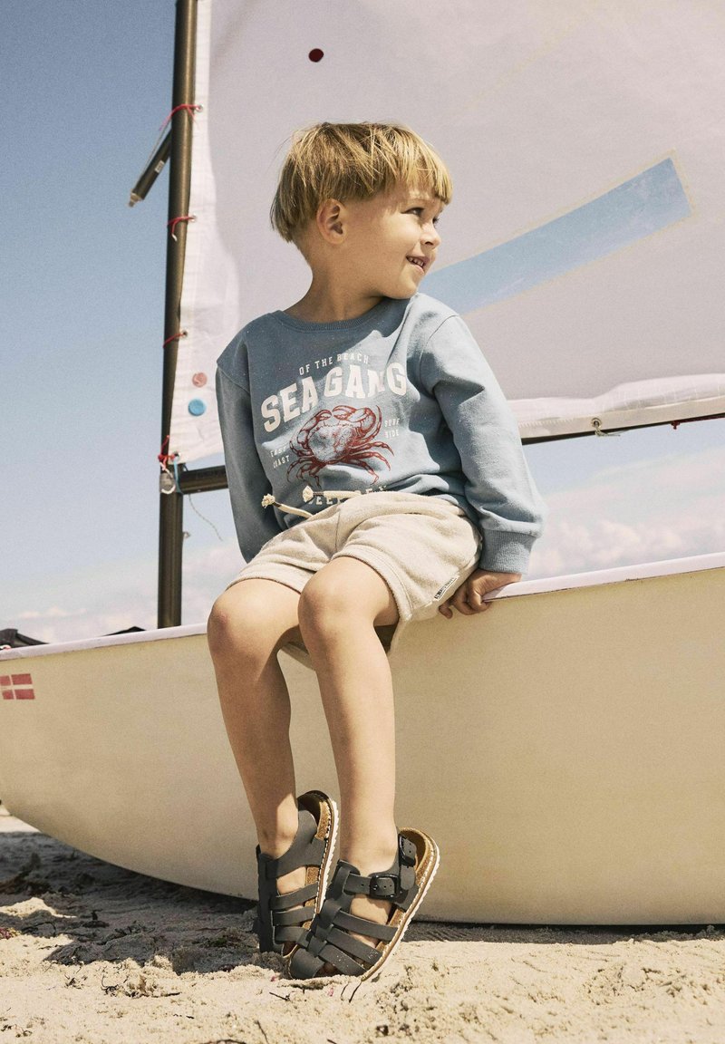 Sandali neri con più cinturini e fibbia regolabile, indossati con pantaloni corti chiari e una felpa blu con grafica che presenta un granchio.