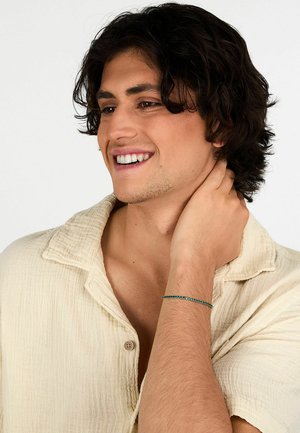 Braccialetto di perline con pietre verdi su un polso. Il portatore ha capelli scuri e indossa una camicia color crema, strutturata, con colletto.