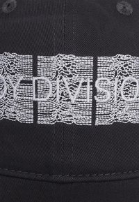 Casquette noire avec le texte brodé en gris "JOY DIVISION" et un motif en dentelle. Le matériel présente une texture typique du tissu en coton.