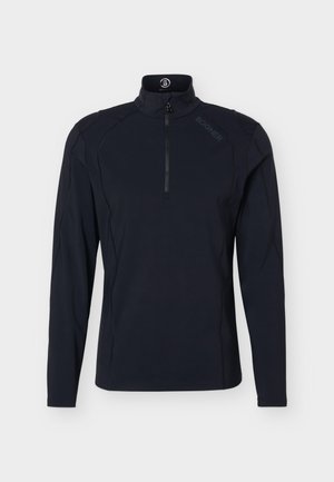Hombre de pie de frente, vistiendo una camiseta negra de manga larga, pantalones beige con cinturón negro y zapatillas blancas sobre un fondo liso.