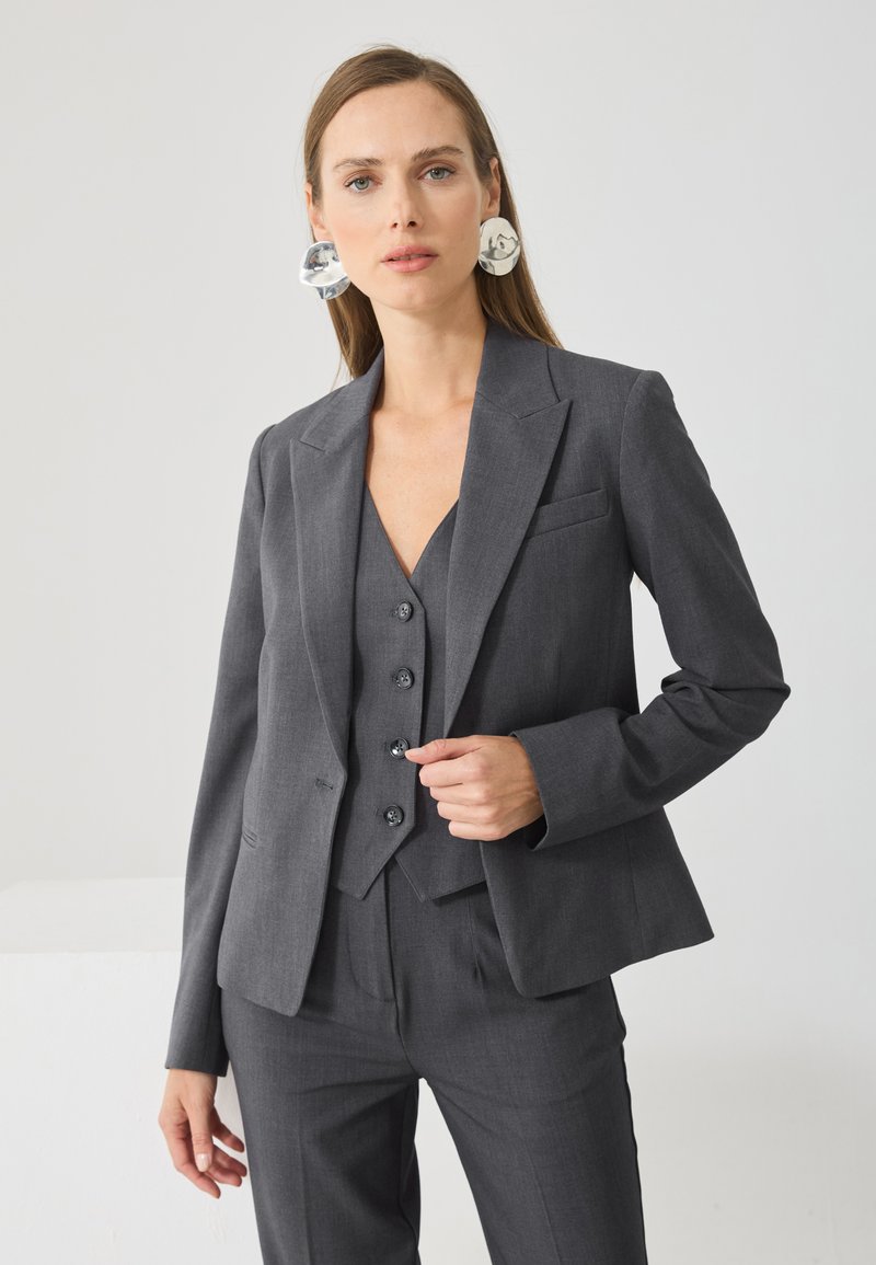 Γκρι κοντομάνικο σακάκι με γιακά notch, κουμπί στο κέντρο και τσέπη στο στήθος, σε συνδυασμό με γκρι γιλέκο που διαθέτει πέντε μαύρα κουμπιά.