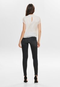 Blusa blanca translúcida con mangas cortas, patrón texturizado, detalle de escote en la espalda, combinada con jeans ajustados negros y zapatos de tacón alto.