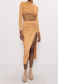 Mujer vistiendo un top corto de manga larga color beige con tirantes envolventes y una falda midi a juego con una gran abertura, combinada con calcetines y zapatos de tobillo beige.