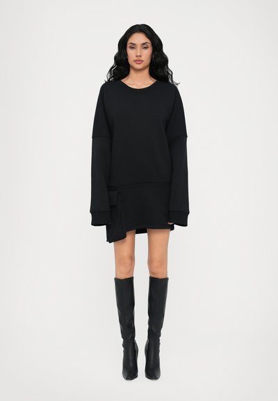 Sort oversized sweatshirtkjole med lange ærmer, rund halsudskæring, sidelomme og asymmetrisk kant, parret med sorte knæhøje støvler.