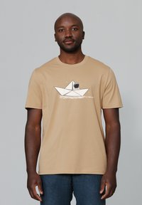 Beige katoenen t-shirt met een geprinte graphic van een papieren boot en een astronaut, met subtiele details van watergolven onder het ontwerp.