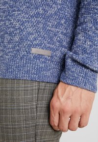 Blauer Strickpullover mit strukturiertem Muster, versehen mit einem Leder-Esprit-Label, kombiniert mit karierter brauner und beiger Hose.