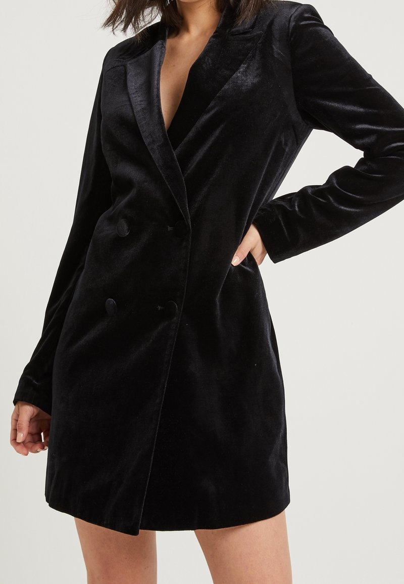 Robe blazer en velours noir avec un revers cranté, des boutons croisés, des manches longues et une coupe cintrée. Texture lisse et longueur au-dessus du genou.