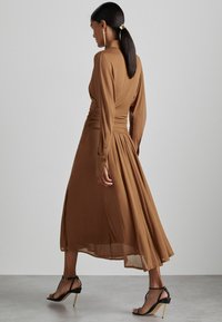 Bruine lange mouwen jurk met gerimpelde taille, vloeiende rok en knoopmanchetten. Draagt een paar zwarte hakken met gouden accent.