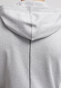 Sudadera gris con capucha ajustable, textura suave, una costura negra central que recorre la parte posterior y bordes sin rematar en la capucha.