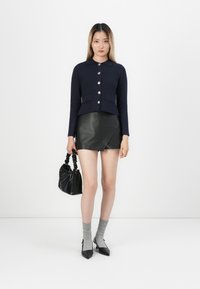 Marineblauwe geknipte brei cardigan met een kraag en gouden knopen, gecombineerd met een zwarte nep-lederen minirok. Model houdt een zwarte handtas vast.