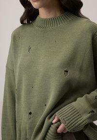 Przesadny zielony sweter dzianinowy z prążkowanym dekoltem i mankietami. Posiada liczne podarte dziury w całym materiale, co tworzy teksturowany wygląd.