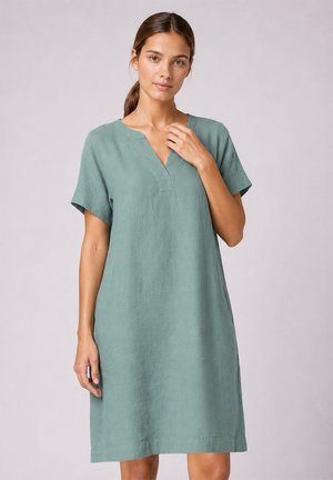 Femme aux cheveux bruns en queue de cheval basse, portant une robe verte ample à manches courtes, longueur genou, avec un col en V.