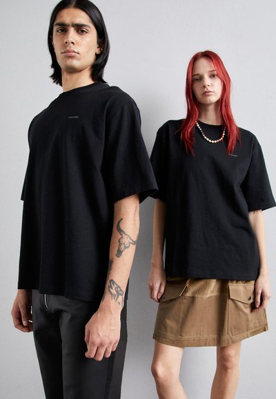 Schwarze, übergroße T-Shirts aus Baumwolle mit kurzen Ärmeln, kombiniert mit einem braunen Cordrock mit großen Taschen und kontrastierenden Nähten.