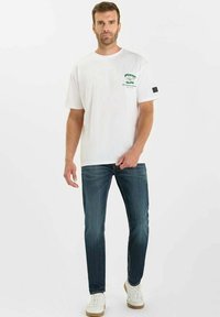 Witte katoenen t-shirt met groen grafisch ontwerp op de linkerkant van de borst, gecombineerd met donkerblauwe spijkerbroek en witte sneakers met lichtbruine zolen.