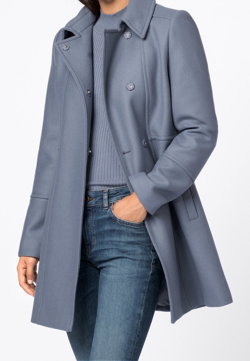 Femme portant un manteau gris à double boutonnage sur un pull côtelé et un jean bleu, avec une main dans la poche, sur un fond uni.