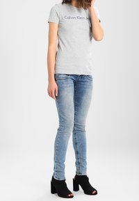 Šedé tričko s krátkým rukávem s nápisem "Calvin Klein Jeans", v kombinaci se světle modrými slim-fit džíny a černými otevřenými vysokými botami na podpatku.