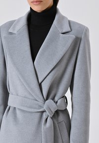Cappotto avvolgente grigio chiaro con tessuto testurizzato, ampi rever e cintura legata. Presenta tasche frontali e una silhouette snella.
