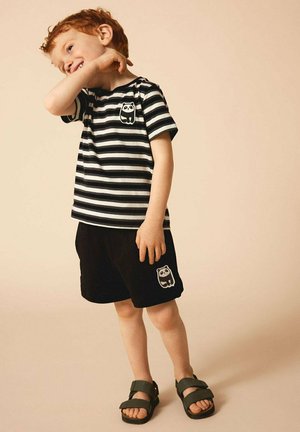 Gestreiftes schwarzes und weißes Baumwoll-T-Shirt mit einem Panda-Grafik, kombiniert mit schwarzen Shorts, die ein ähnliches Panda-Motiv aufweisen, und grünen Sandalen.