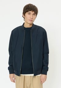 Jeune homme portant une veste bomber zippée bleu marine sur un pull foncé et un pantalon beige, sur un fond blanc uni.