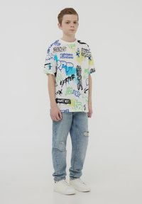 T-shirt grafica multicolore con vari testi e design, abbinata a jeans chiari strappati e sneakers bianche.