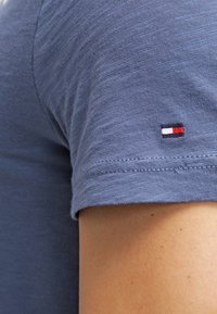 Camisa azul de manga corta con un pequeño logotipo bordado en rojo, blanco y azul cerca del dobladillo, sobre piel clara.