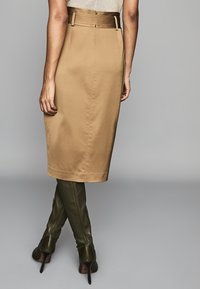 Vrouw in een hoog-gewijze beige kokerrok met een bijpassende riem en donkergroene kniehoogte hakken, staand tegen een grijze achtergrond.