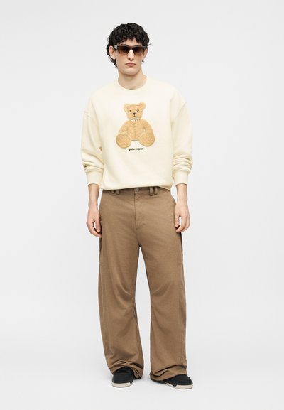 Άνδρας με γυαλιά ηλίου, μπεζ φούτερ με σχέδιο αρκουδάκι, άνετο καφέ παντελόνι και μαύρα αθλητικά παπούτσια, στέκεται μπροστά σε λευκό φόντο.