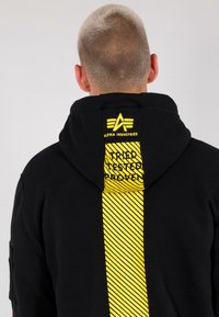 Persona con capelli biondi corti che indossa un hoodie nero con un design a strisce gialle verticali e il testo "Alpha Industries" sul cappuccio.