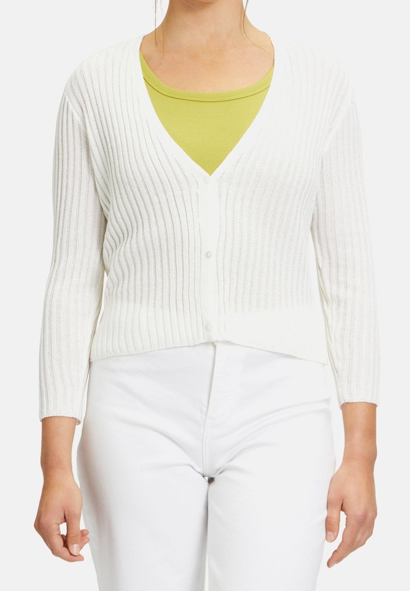 Bílý žebrovaný cardigan s výstřihem do V, s knoflíkovým zapínáním a tříčtvrtečními rukávy, přeložený přes zelený top. Textura vypadá měkce a elasticky.