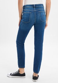 Dunkelblaue Denim-Jeans mit taillierter Passform, zwei Gesäßtaschen und weißen Nähten. Kombiniert mit schwarzen Sneakers mit niedriger Schafthöhe.
