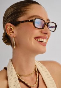 Νεαρή γυναίκα που χαμογελά, φορά γυαλιά με όψη χελώνας, χρυσό σπιράλ σκουλαρίκι στο αυτί, κρίκο και πολλαπλά στρωμένα κολιέ με χάντρες, μαζί με ένα κρεμ πουλόβερ πλέξης.