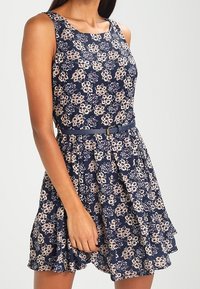 Marint blommig klänning med ärmlös design och vågig nederkant. Har en bälte i midjan och ett mönster av ljusrosa och krämfärgade blommor.