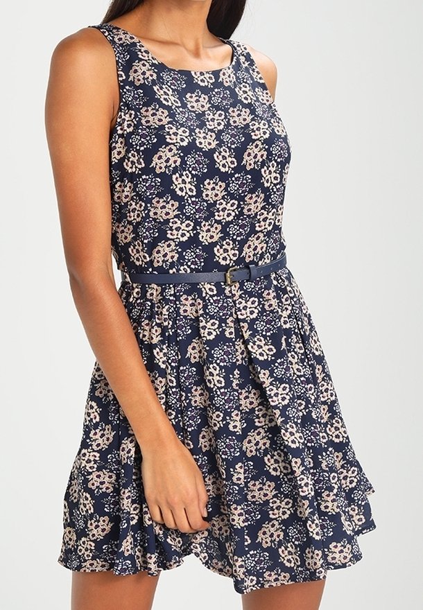 Marint blommig klänning med ärmlös design och vågig nederkant. Har en bälte i midjan och ett mönster av ljusrosa och krämfärgade blommor.