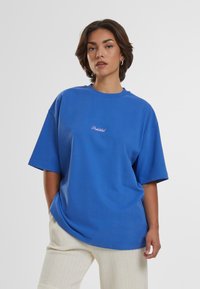 Blå oversized t-skjorte laget av bomull, med korte ermer og rund hals. Sentralisert brodert tekst "Forbudt" i hvitt.