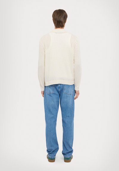 Cream ribbed knit cardigan with a collar, featuring long sleeves and an open back; paired with light blue straight-leg jeans.