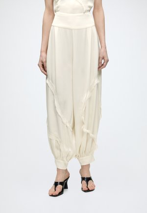 Femme portant un pantalon large couleur crème avec des coutures diagonales à franges et des chevilles retroussées, associé à des sandales noires à talons hauts.