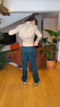 Μπεζ πουκάμισο με κουμπιά και σκούρο πράσινο παντελόνι. Βαμβακερό υλικό, εφαρμοστό σχήμα. Εσωτερικός χώρος με καναπέ και φυτευτό φυτό. Φυσικό ξύλινο πάτωμα.