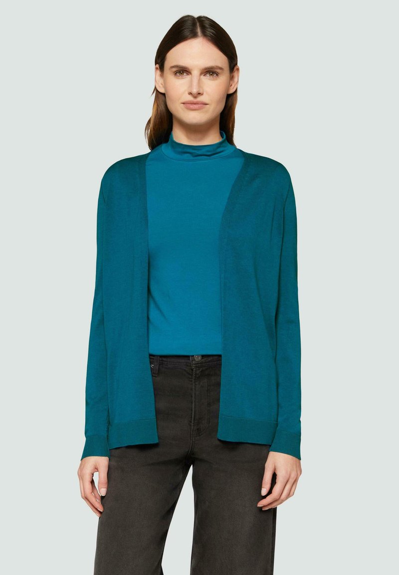 Teal Cardigan mit offenen Vorderseite, aus weichem Stoff, lange Ärmel und gerippte Bündchen. Kombiniert mit einem passenden tealfarbenen Rollkragenpullover und dunklen Hosen.