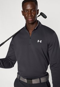 Μαύρο αθλητικό πούλoβερ με φερμουάρ και λευκό λογότυπο, σε συνδυασμό με γκρι ζώνη με υφή· γκολφ ρόπαλο κρατημένο πάνω στον ώμο.