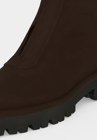 Gros plan d'une botte marron foncé avec une tige lisse et une semelle épaisse en caoutchouc noir avec des crampons profonds pour l'adhérence.